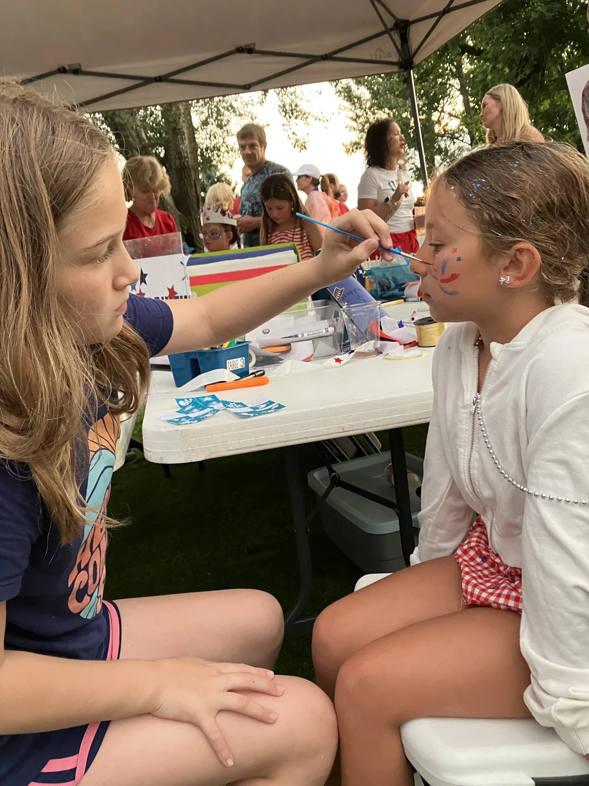 A teen volunteer painting a red, white, and blue star on a young girl's nose at the face painting tent