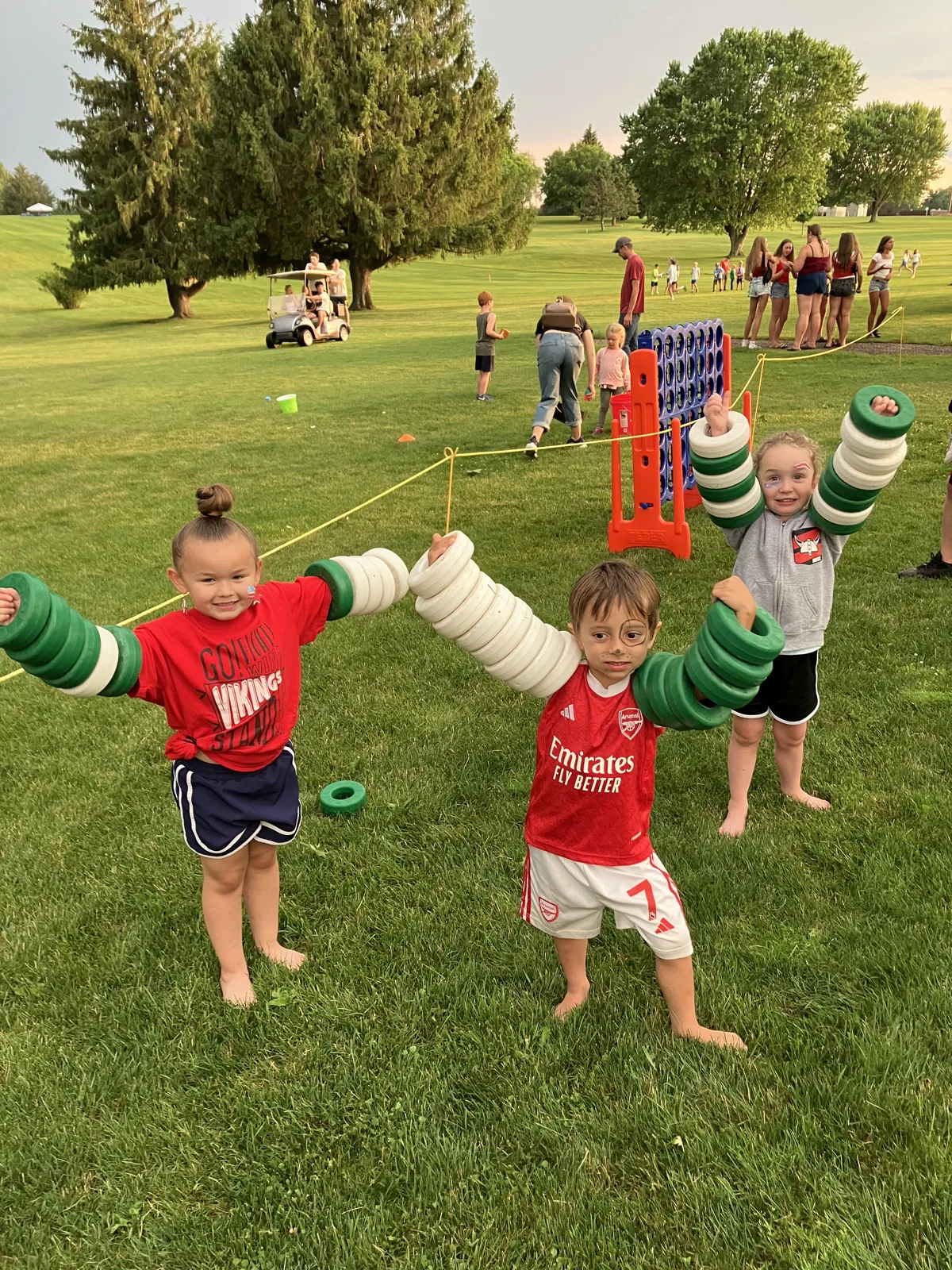 Three kids grinning with ring-toss rings stacked up their arms like muscles on the golf course lawn