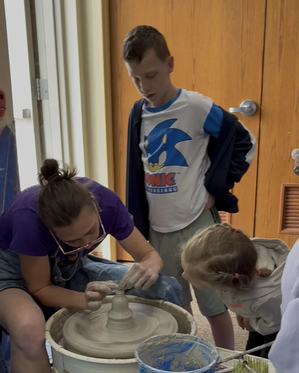 A potter throwing on a demonstration wheel while two young visitors lean in to watch