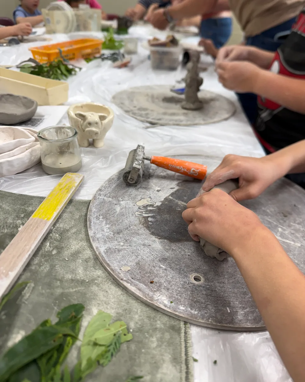 Hands shaping clay at a hand-building station, with rollers, tools, and a small clay pig on the table