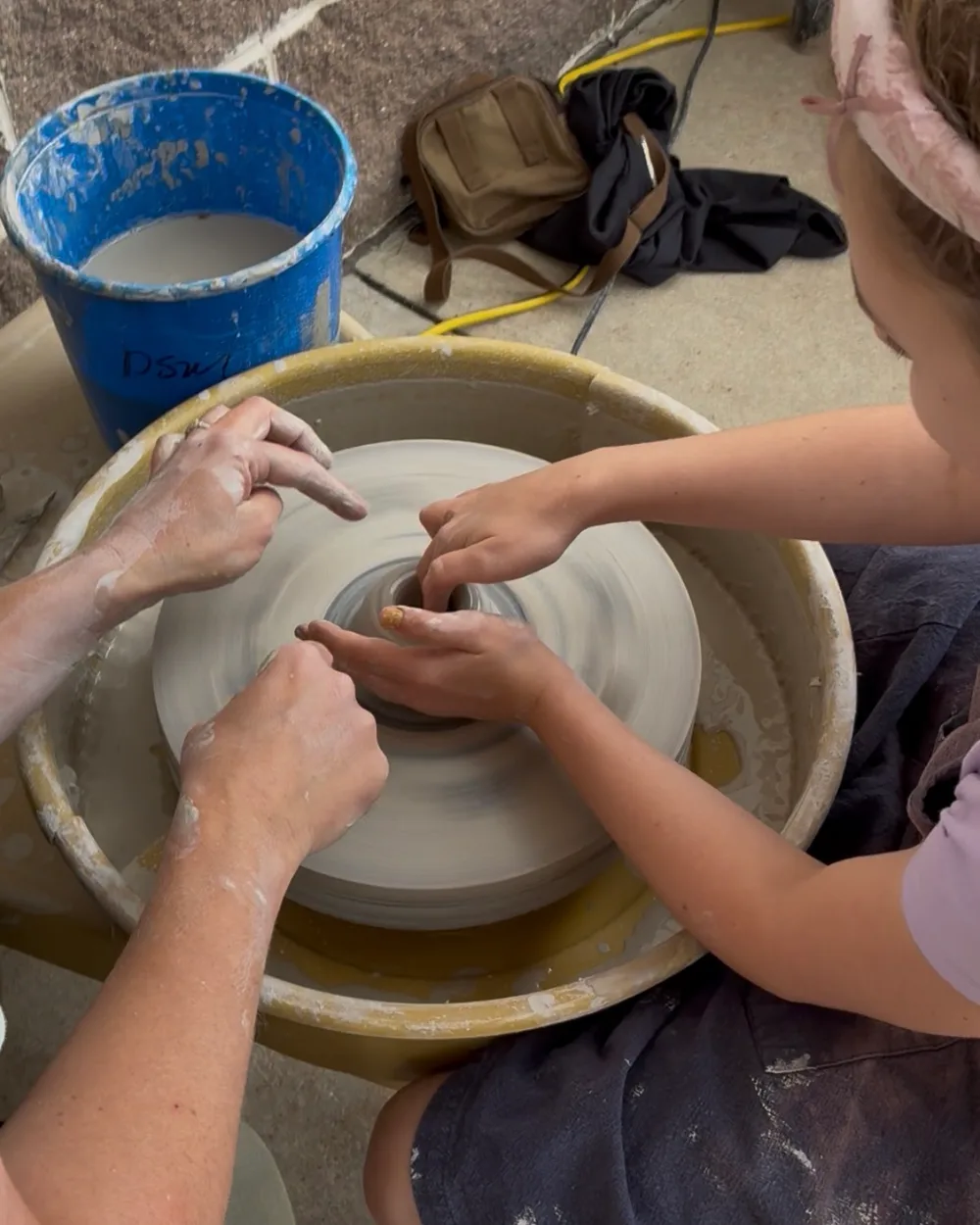 A potter guiding a young visitor's hands to center clay on the wheel